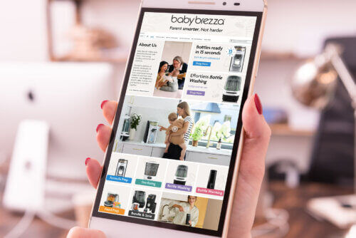 Cheeky Rascals Baby Brezza Amazon listings - Venture Forge Tablet displaying the Baby Brezza website held by a person with red nail polish, showing product tiles and 'About Us' section on a kitchen-like page.