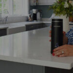 Cheeky-overlay - Venture Forge Person with red nails rests a black insulated tumbler on a white kitchen island.