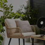Glen Dimplex overlay - Venture Forge Outdoor patio scene with wicker chairs, beige cushions, and lush green plants, plus a black pedestal fan on a small table nearby.