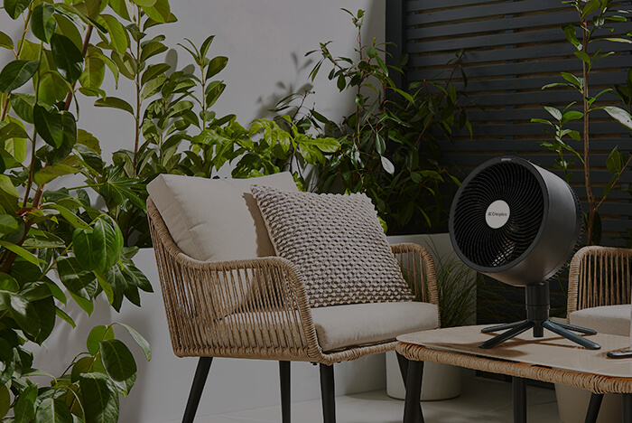 Glen Dimplex overlay - Venture Forge Outdoor patio scene with wicker chairs, beige cushions, and lush green plants, plus a black pedestal fan on a small table nearby.