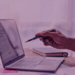 Why-Amazon-Peak-Events-expose-the-Brands-that-werent-ready-Venture-Forge - Venture Forge Person's hand holds a pen over an open laptop on a wooden desk, with notebook and papers nearby, in a business setting.