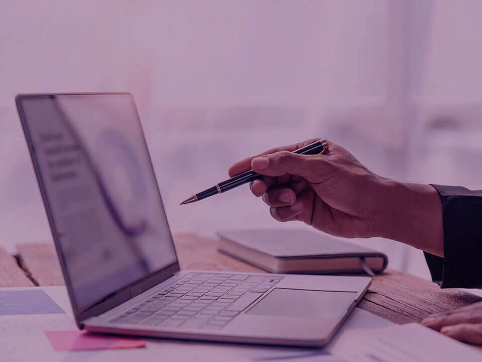 Why-Amazon-Peak-Events-expose-the-Brands-that-werent-ready-Venture-Forge - Venture Forge Person's hand holds a pen over an open laptop on a wooden desk, with notebook and papers nearby, in a business setting.