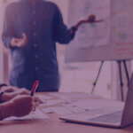 Are you blaming your Amazon Ads for a Listings Problem From Amazon Agency Venture Forge - Venture Forge Team meeting: presenter gestures toward a whiteboard while others write notes and a laptop sits on the table.