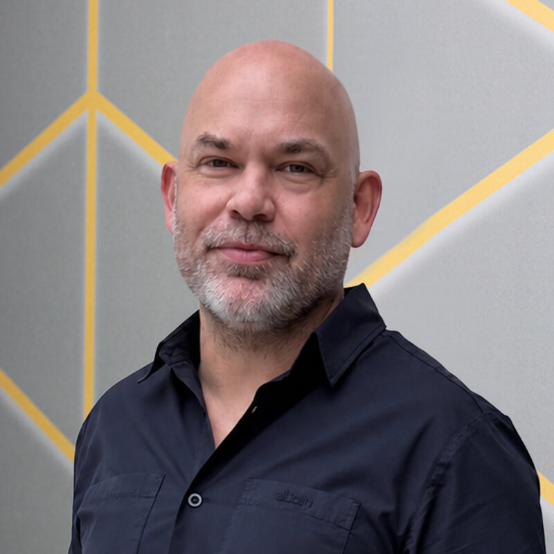 Joel Turner Head of Growth Commercial Director Venture Forge - Venture Forge Portrait of a smiling bald man with a gray beard, wearing a dark shirt, against a gray wall with yellow geometric lines.