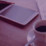 Hand resting on a white tablet on a wooden desk beside a cup of coffee and a newspaper (purple lighting).