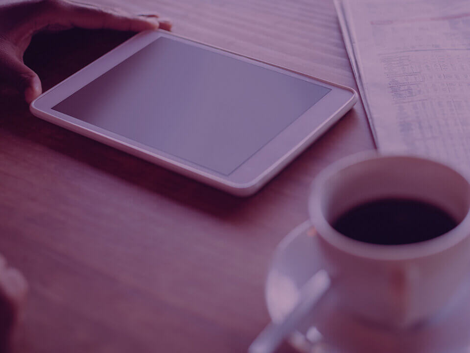 Hand resting on a white tablet on a wooden desk beside a cup of coffee and a newspaper (purple lighting).