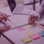 The Mid-Year Amazon Numbers Most Brands Read the Wrong Way by Venture Forge Amazon Agency - Venture Forge Two professionals review a document on a desk, pointing with pens over charts and post-it notes. The scene includes a laptop and sticky notes nearby.