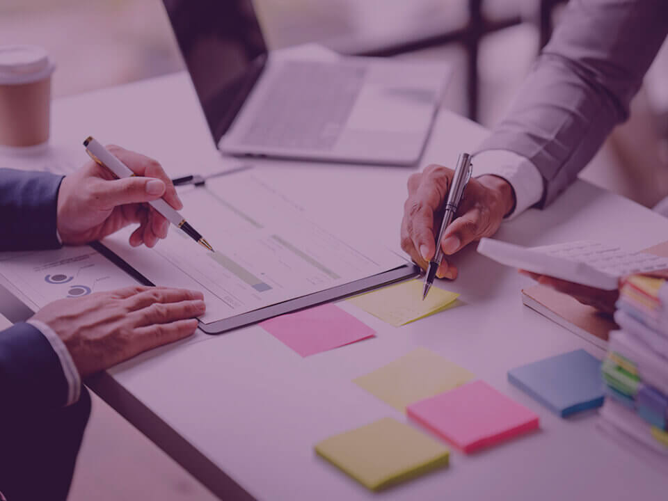 The Mid-Year Amazon Numbers Most Brands Read the Wrong Way by Venture Forge Amazon Agency - Venture Forge Two professionals review a document on a desk, pointing with pens over charts and post-it notes. The scene includes a laptop and sticky notes nearby.