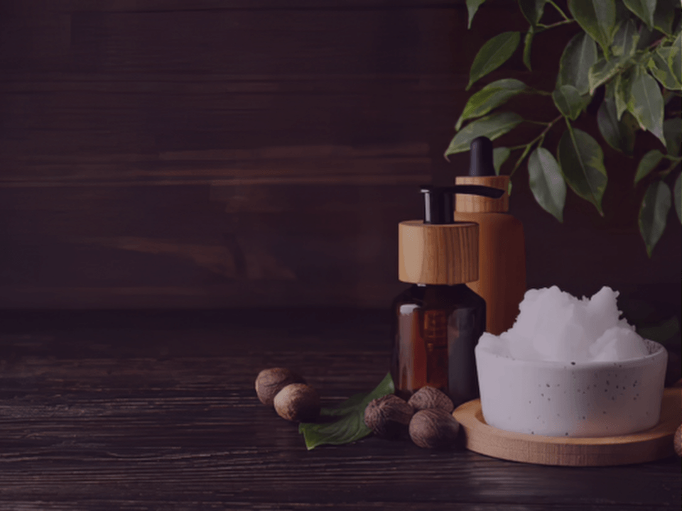 Natural spa scene with a white bowl of bath salts, amber bottles with wooden tops, walnuts, and green leaves on a dark wooden surface.